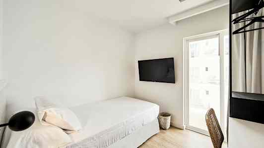 Hotellihuone, jossa on yhden hengen sänky, seinälle kiinnitetty TV ja työpöytä tuoleineen ikkunan vieressä. (Tekoälyn luoma teksti)