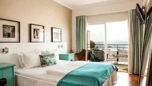 Hotellihuone, jossa on parivuode ja valkoiset ja siniset vuodevaatteet, seinätaidetta sängyn yläpuolella ja parveke merinäköalalla. (Tekoälyn luoma teksti)