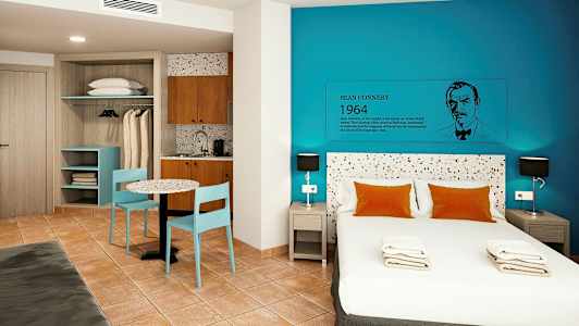 Moderni hotellihuone, jossa on parisänky, siniset seinät ja pieni keittiönurkkaus. (Tekoälyn luoma teksti)