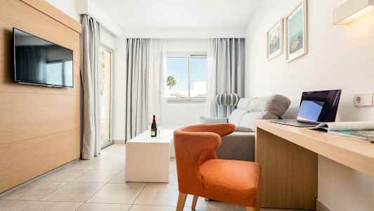 Valoisa, moderni hotellihuone, jossa on sohva, tuoli, televisio, kirjoituspöytä ja suuret ikkunat. (Tekoälyn luoma teksti)