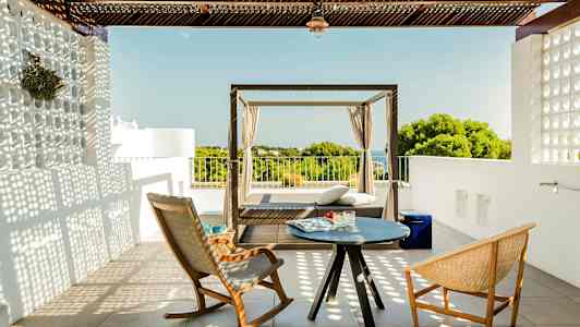 Terassi, jossa kaksi tuolia ja pieni pöytä sekä päivänsänky näkymällä vehreyteen ja merelle. (Tekoälyn luoma teksti)