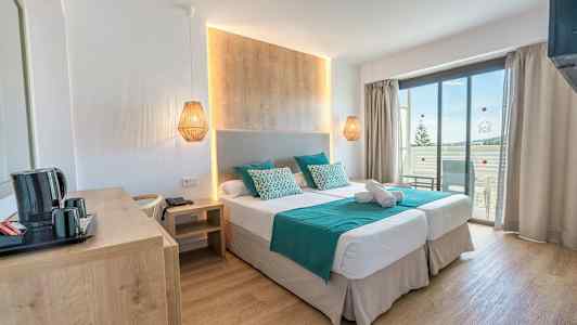 Hotellihuone, jossa kaksi erillistä vuodetta, puupaneli sängyn takana, turkoosit lakanat ja tyynyt sekä parveke näköalalla. (Tekoälyn luoma teksti)