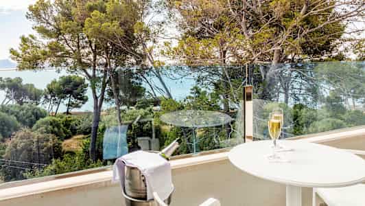 Parveke, jolta on näkymä puihin ja merelle. Parvekkeella on valkoinen pöytä, jonka päällä on samppanjalasi, sekä teline, jossa on jäähdytysastia samppanjapullolle. (Tekoälyn luoma teksti)