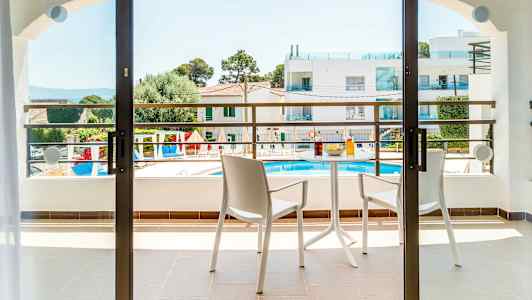 Parveke, jossa kaksi tuolia ja pöytä, jossa juomia ja välipalaa. Näkymä allasalueelle ja ympäröiviin rakennuksiin. (Tekoälyn luoma teksti)