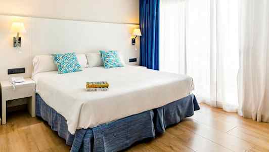 Hotellihuone, jossa on leveä sänky, valkoinen sängynpääty, siniset tyynyt ja verhot sekä vaalea puulattia. (Tekoälyn luoma teksti)