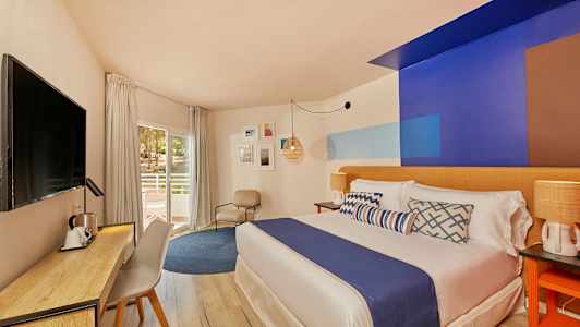 Kuva hotellihuoneesta, jossa on moderni sisustus, sänky sinisellä päiväpeitteellä, kirjoituspöytä ja tuoli sekä parvekkeen ovi. (Tekoälyn luoma teksti)