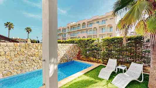 Kuva ulkouima-altaasta, jota ympäröivät kiviseinät ja vehreä puutarha, kaksi valkoista aurinkotuolia ja taustalla kaunis hotelli. (Tekoälyn luoma teksti)