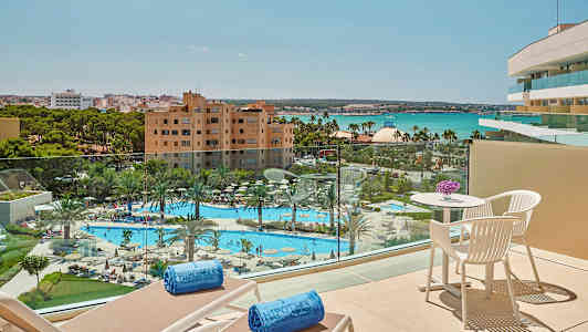 Näkymä parvekkeelta uima-altaalle ja puutarhaan, joissa on palmuja, hotellirakennuksia ja merta taustalla. (Tekoälyn luoma teksti)