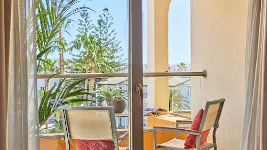 Näkymä parvekkeen läpi, jossa on kaksi tuolia ja pöytä, ympäröivät kasvit ja rakennukset. (Tekoälyn luoma teksti)