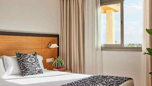 Hotellihuone, jossa on sänky, yöpöytä ja ikkuna, joka päästää luonnonvaloa sisään. (Tekoälyn luoma teksti)