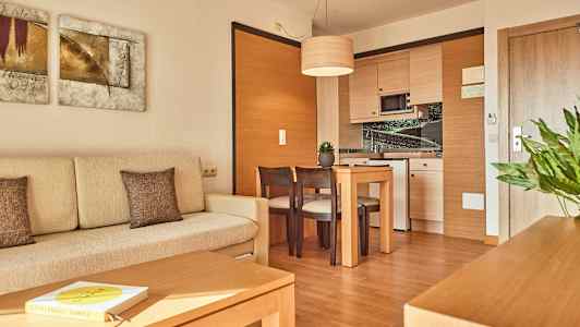 Valokuva modernista hotellihuoneesta, jossa on vaalea sohva, puinen pöytä tuoleineen ja nurkkakeittiö. (Tekoälyn luoma teksti)