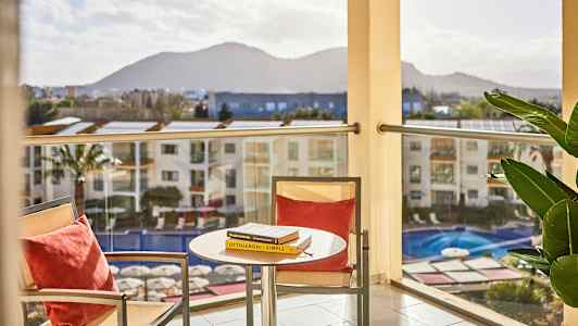 Näkymä parvekkeelta, jossa etualalla on pöytä ja kaksi tuolia, sekä alla oleva uima-allas. Taustalla näkyy vuoria. (Tekoälyn luoma teksti)