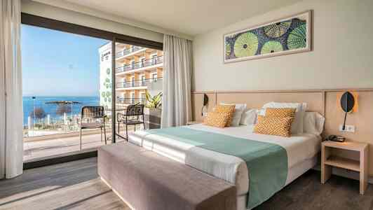 Hotellihuone, jossa sänky, istuinpaikat ja näkymä merelle suurten ikkunoiden läpi. (Tekoälyn luoma teksti)