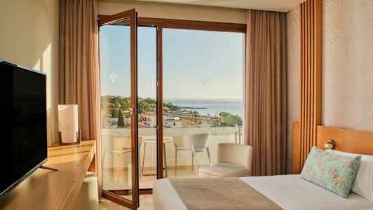 Hotellihuone, jossa on sänky, työpöytä, televisio ja avoin parvekkeen ovi, joka johtaa parvekkeelle merinäköalalla ja puiden ympäröimänä. (Tekoälyn luoma teksti)