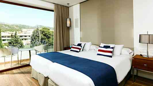 Hotellihuone, jossa on suuri parisänky, sinivalkoiset vuodevaatteet ja ikkuna näkymällä vehreyteen ja rakennuksiin. (Tekoälyn luoma teksti)