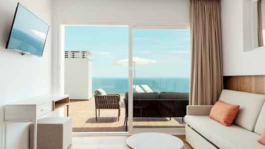 Valoisa huone, jossa on sohva ja pöytä, ulkoterassilla istuimia ja näkymä merelle. (Tekoälyn luoma teksti)