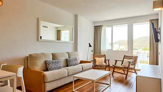 Olohuone, jossa on beige sohva, kaksi tuolia ja valkoinen sohvapöytä. Ikkunasta näkyvät vehreät maisemat. (Tekoälyn luoma teksti)