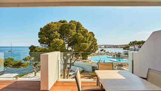 Näköala ulkoterassille, jossa on pöytiä ja tuoleja, ulkouima-allasalueelle ja merelle, jossa etäisyydessä purjevene. (Tekoälyn luoma teksti)