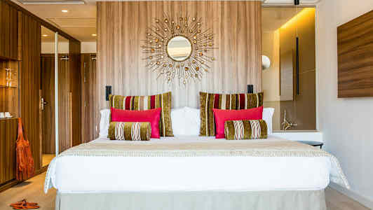 Moderni hotellihuone, jossa on suuri sänky, koristeellisia tyynyjä eri väreissä ja takaseinällä puupaneelia. (Tekoälyn luoma teksti)