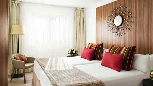 Moderni hotellihuone, jossa kaksi vuodetta, värikkäitä tyynyjä ja minimalistinen sisustus. (Tekoälyn luoma teksti)