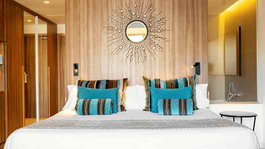 Hotellihuone, jossa suuri sänky koristeltuna värikkäillä tyynyillä sinisenä ja raidallisena kuviona, puuseinä peilimotiivilla ja moderni valaistus. (Tekoälyn luoma teksti)