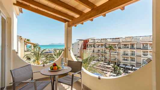 Parveke, jossa kaksi tuolia ja pöytä hedelmäkulholla, merinäköala ja hotellirakennukset taustalla. (Tekoälyn luoma teksti)