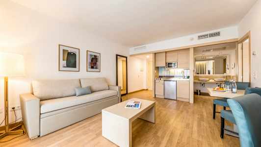 Nykyaikainen hotellihuone, jossa beige sohva ja pöytä puulattialla sekä keittiöosa ja kylpyhuone taustalla. (Tekoälyn luoma teksti)