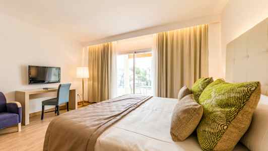 Hotellihuone, jossa on suuri sänky, vihreät tyynyt, pöytä, tuoli ja televisio. Ikkunat verhoilla päästävät luonnonvaloa sisään. (Tekoälyn luoma teksti)