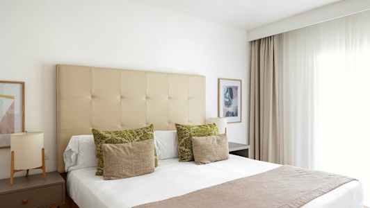 Valoisa ja tyylikäs hotellihuone, jossa on pedattu sänky. Verho oikealla ja yöpöytä vasemmalla. (Tekoälyn luoma teksti)