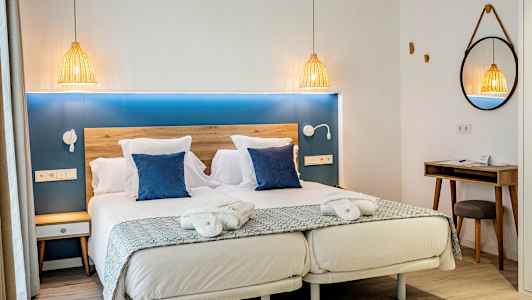 Hotellihuone, jossa on parivuode, kaksi sinistä tyynyä, yöpöytä ja peili. (Tekoälyn luoma teksti)