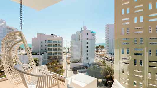 Parveke, jossa on valkoiset tuolit ja näkymä moderneihin rakennuksiin sekä taustalla olevaan mereen. (Tekoälyn luoma teksti)