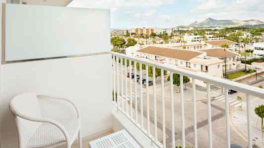 Parveke, jossa on kaksi valkoista tuolia ja pöytä, näkymä kadulle ja rakennuksiin. (Tekoälyn luoma teksti)