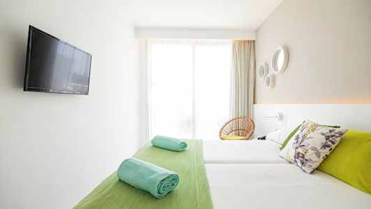 Valoisa hotellihuone, jossa on parivuode, värikkäitä tyynyjä, rullattuja pyyhkeitä ja seinälle asennettu taulutelevisio. (Tekoälyn luoma teksti)