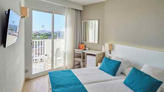 Valoisa hotellihuone, jossa kaksi sänkyä, siniset tyynyt ja päiväpeitteet sekä parveke näkymällä. (Tekoälyn luoma teksti)