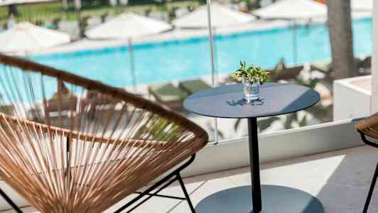 Istuinryhmä, jossa pöytä ja tuolit, ikkunan edessä näkyy uima-allas ja aurinkotuoleja. (Tekoälyn luoma teksti)