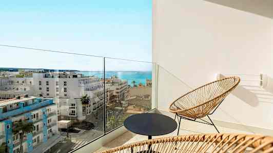 Parveke, jossa on kaksi rottinkituolia ja pyöreä pöytä, näkymä merelle ja kaupungin rakennuksiin. (Tekoälyn luoma teksti)