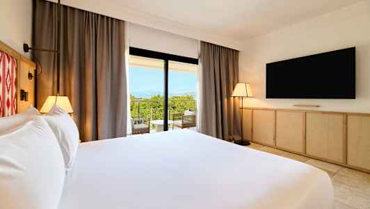 Hotellihuone, jossa iso sänky, televisio seinällä ja parveke, josta näkymä vehreään maisemaan. (Tekoälyn luoma teksti)