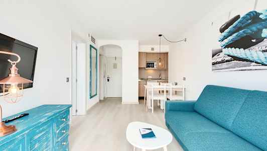 Valoisa hotellihuone, jossa on turkoosi teema, sisältäen sohvan, pöydän, television ja keittokomeron. (Tekoälyn luoma teksti)