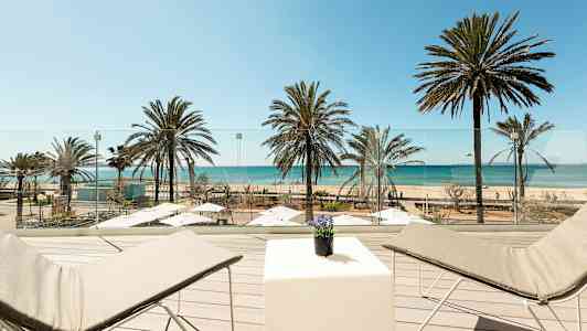 Terassin näkymä, jossa aurinkotuoleja ja pöytä, ranta palmuineen ja meri. (Tekoälyn luoma teksti)