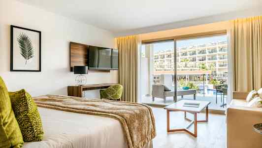 Hotellihuone, jossa on sänky, sohvaryhmä ja parveke näkymällä uima-allasalueelle ja hotellille. (Tekoälyn luoma teksti)