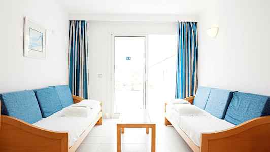 Kaksi sinisillä tyynyillä varustettua sohvaa on vastakkain, välissä puurunkoinen pöytä, taustalla sinivalkoiset verhot ja ikkuna. (Tekoälyn luoma teksti)