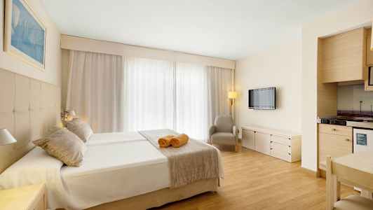 Hotellihuone, jossa on parivuode, kaksi pyyhettä sängyllä, nojatuoli, seinälle kiinnitetty televisio ja pieni keittonurkkaus. (Tekoälyn luoma teksti)