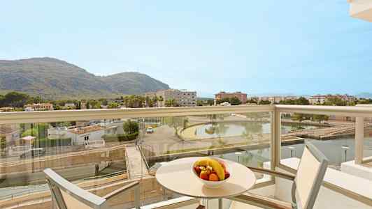 Parveke, jolta on näkymä maisemaan, joka sisältää vuoria, rakennuksia ja lammen. Pöydällä on kulhollinen hedelmiä. (Tekoälyn luoma teksti)