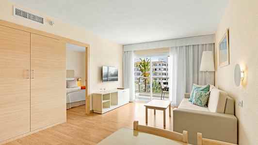 Hotellihuone, jossa on olohuone, sohva tyynyineen, pöytä, televisio ja parveke näkymällä. (Tekoälyn luoma teksti)