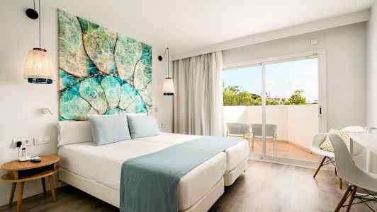 Hotellihuone, jossa on parisänky, siniset vuodevaatteet, värikäs taulu sängyn yläpuolella ja parveke, jonka ulkopuolella on kaksi tuolia. (Tekoälyn luoma teksti)