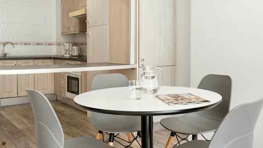 Ruokailutila, jossa pyöreä pöytä, harmaat tuolit ja keittiötila, jossa on puiset kaapit ja kodinkoneet. (Tekoälyn luoma teksti)