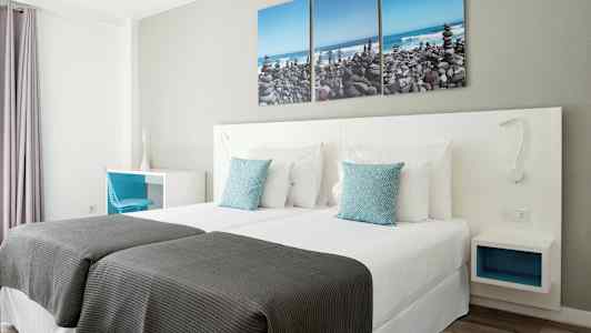 Hotellihuone, jossa on kaksi yksittäissänkyä, valkoiset vuodevaatteet ja siniset koristeelliset tyynyt. Seinällä on kolmen osan taulu, joka esittää kivirantaa. (Tekoälyn luoma teksti)