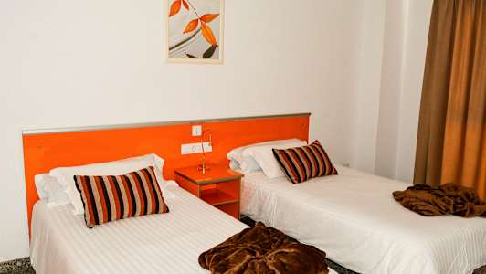 Kaksi yhden hengen sänkyä, joissa on valkoiset lakanat, ruskeat peitot ja raidalliset tyynyt, huoneessa, jossa on oranssi sisustus. (Tekoälyn luoma teksti)