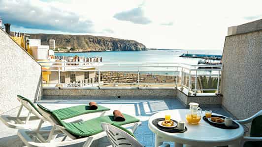 Terassi, jossa kaksi lepotuolia ja pöytä, jossa appelsiinimehua ja leivonnaisia, merenäköala taustalla. (Tekoälyn luoma teksti)