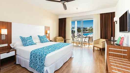 Hotellihuone, jossa suuri sänky, siniset koristeelliset tyynyt ja peite sekä pöytä tuoleineen lasioven vieressä, joka johtaa parvekkeelle. (Tekoälyn luoma teksti)